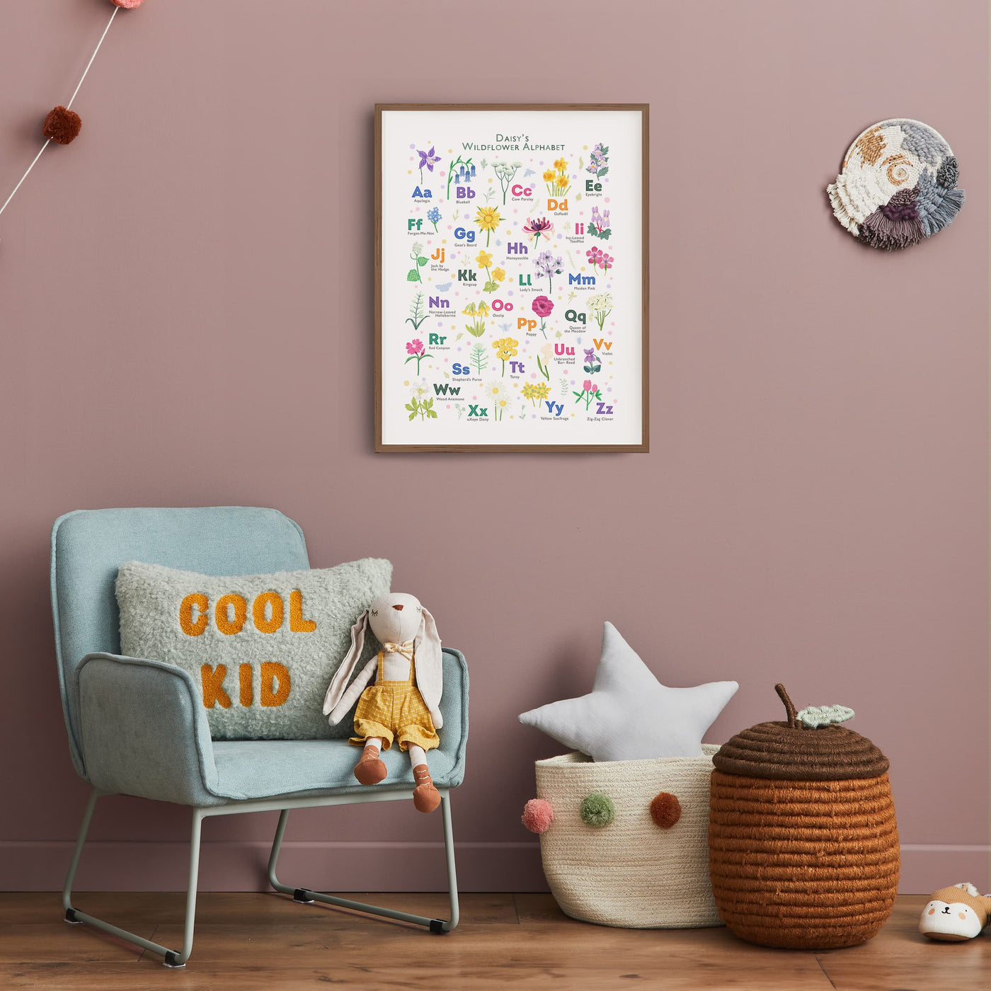 Image of a framed personalized wildflower alphabet print hanging on a dusky pink wall in a child's room