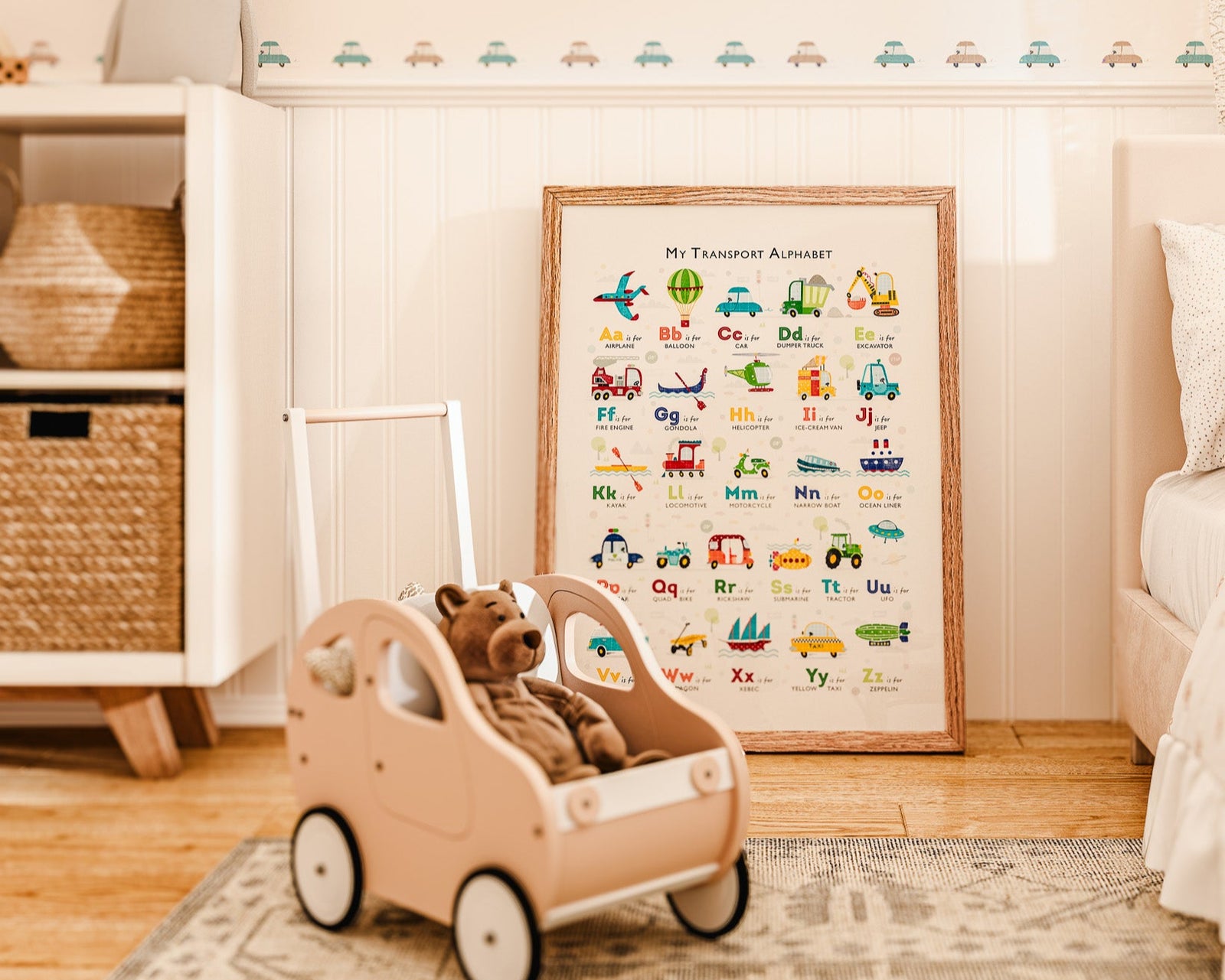 Children's room with teddy bear in a toy car, framed vehicle alphabet print, and car-themed wallpaper.