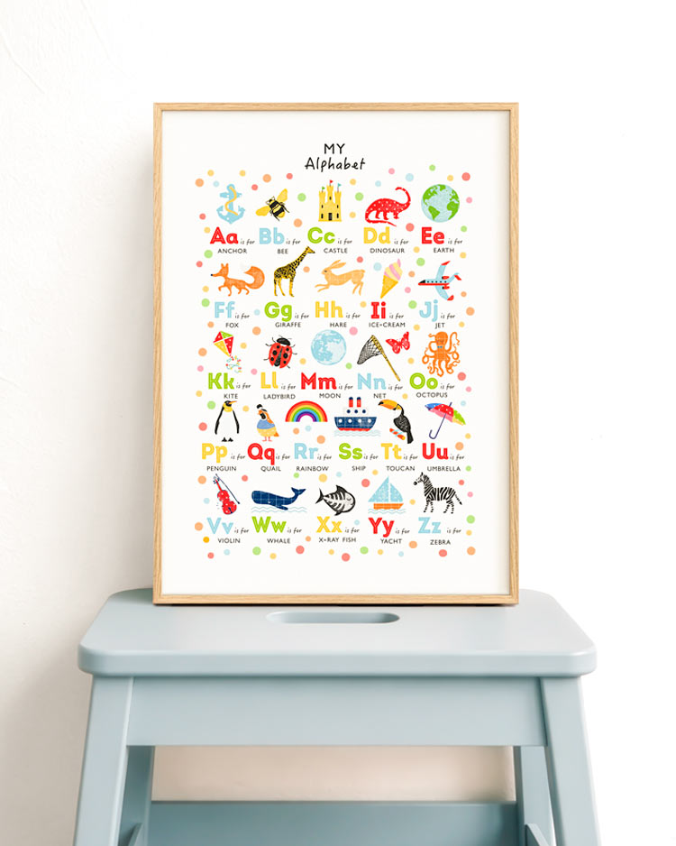 A framed children's alphabet art print leaning on a light blue stool against a white wall. The print features colorful uppercase and lowercase letters with corresponding illustrations such as anchors, elephants, giraffes, and rainbows, under the title 'MY Alphabet'.