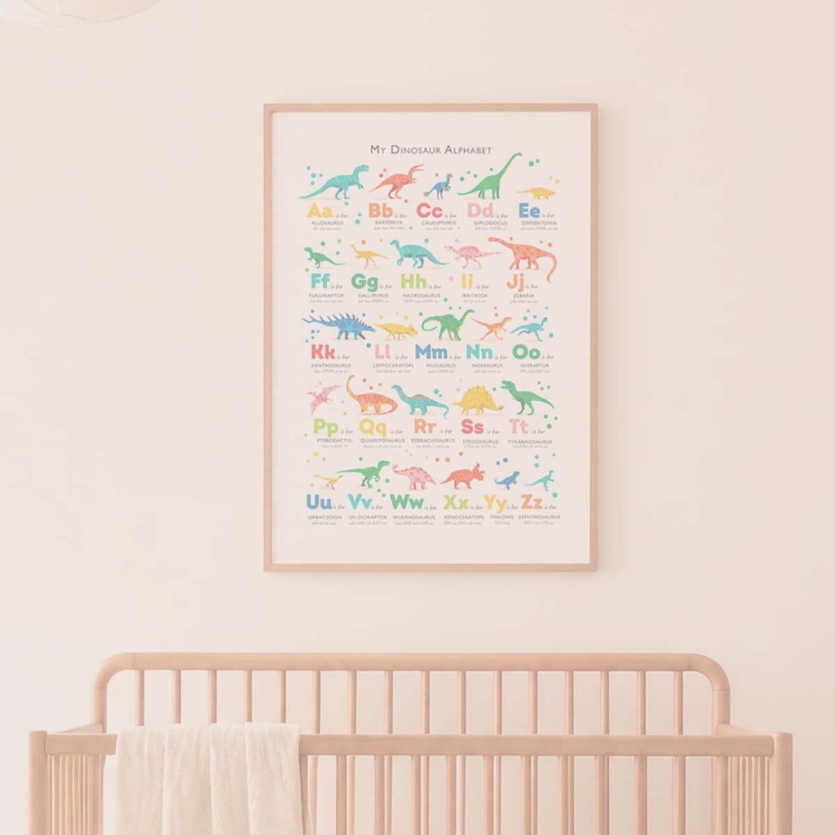 A framed 'Mr. Dinosaur Alphabet' nursery print hanging on a warm beige wall above a light wood crib.