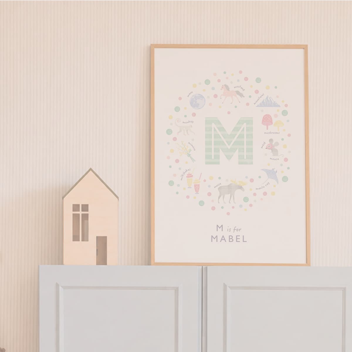 A framed personalized children's wall art print featuring a pastel circular illustration around a large letter M, with the text 'M is for MABEL' at the bottom, displayed on a light wall above a white cabinet next to a small wooden house decoration.