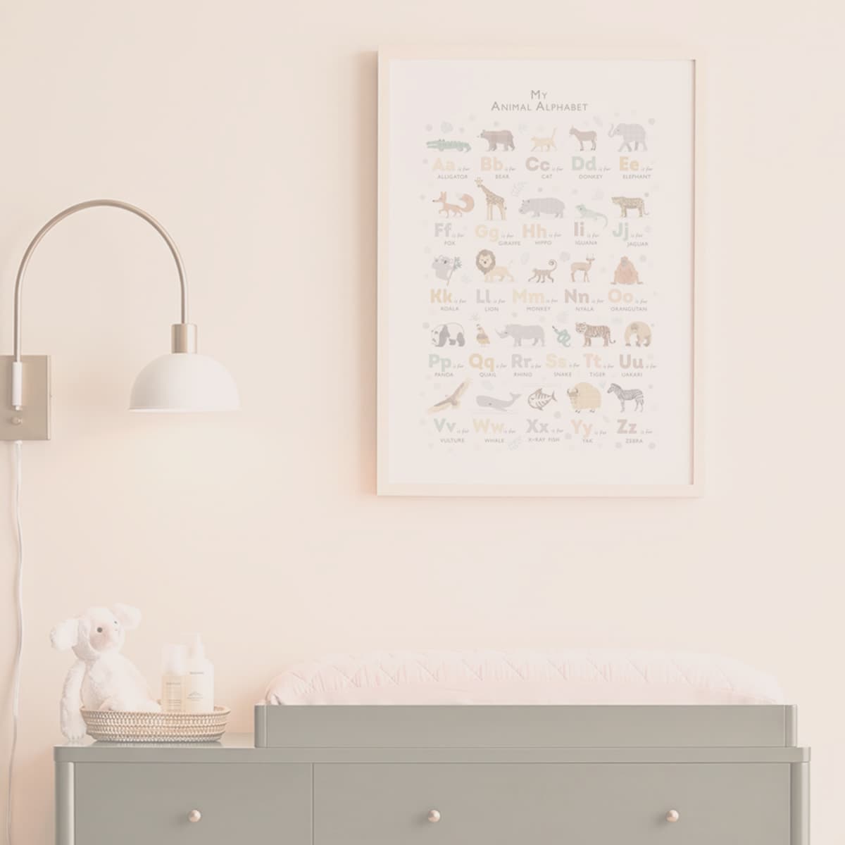 A framed 'My Animal Alphabet' nursery art print hanging on a light blush pink wall above a gray changing table, with a white wall lamp on the left.
