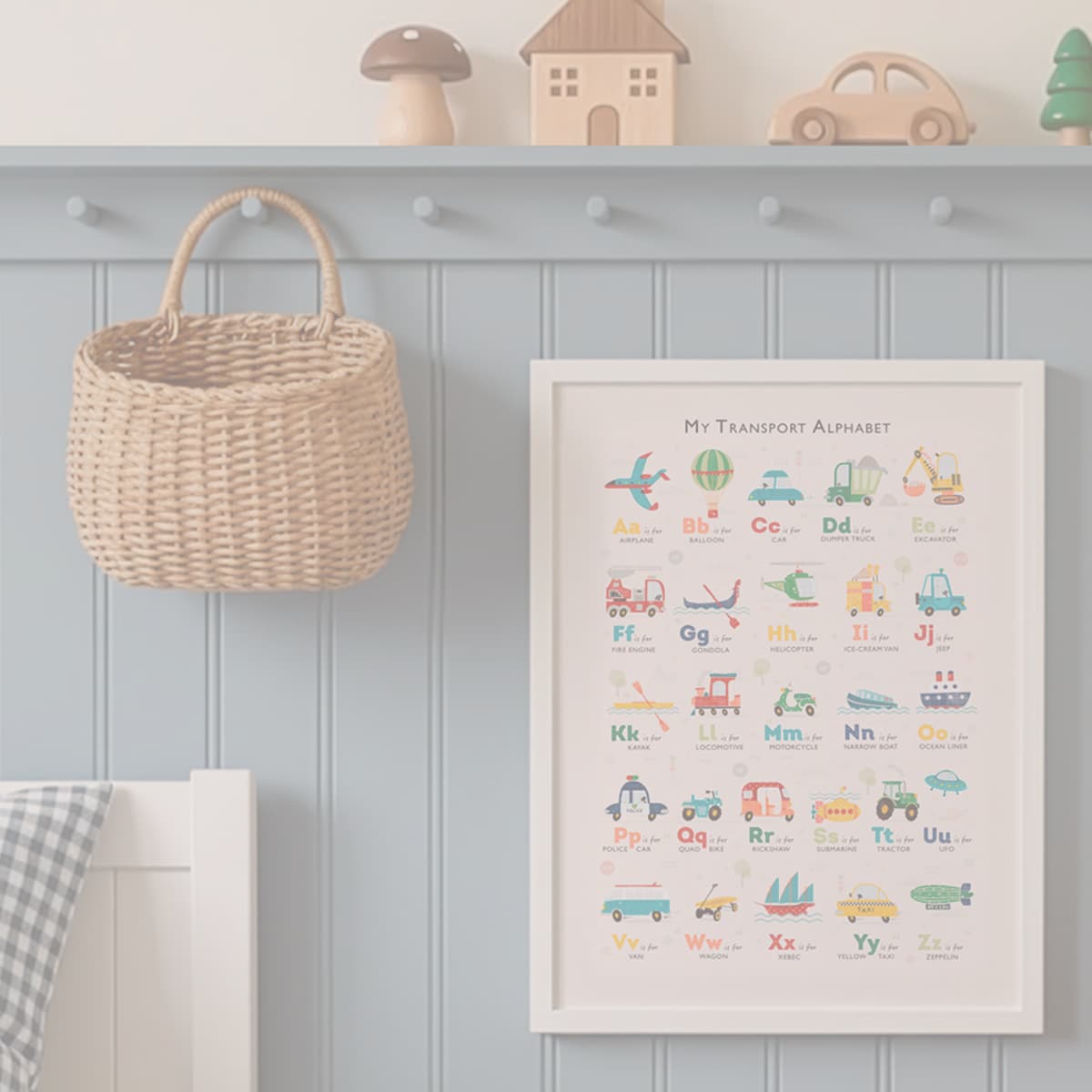 A framed children's educational poster titled 'My Transport Alphabet' hangs on a blue paneled wall in a nursery setting, next to a woven basket and below a shelf with wooden toys.