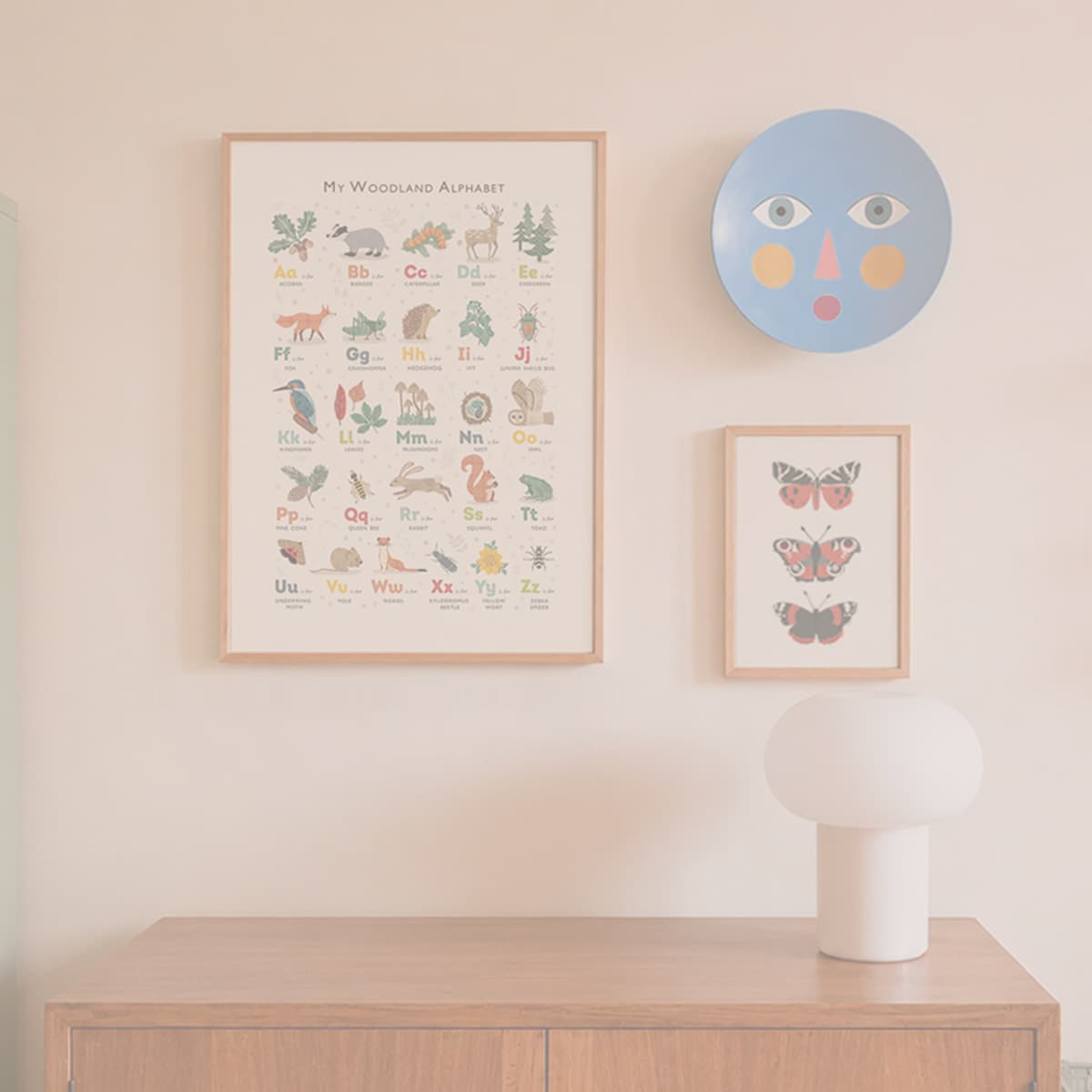 A styled interior scene showing framed wall art hanging above a light wood console table. The main product appears to be the framed alphabet poster reading "My Woodland Alphabet," with additional decor including a blue circular wall plaque and a smaller butterfly print.