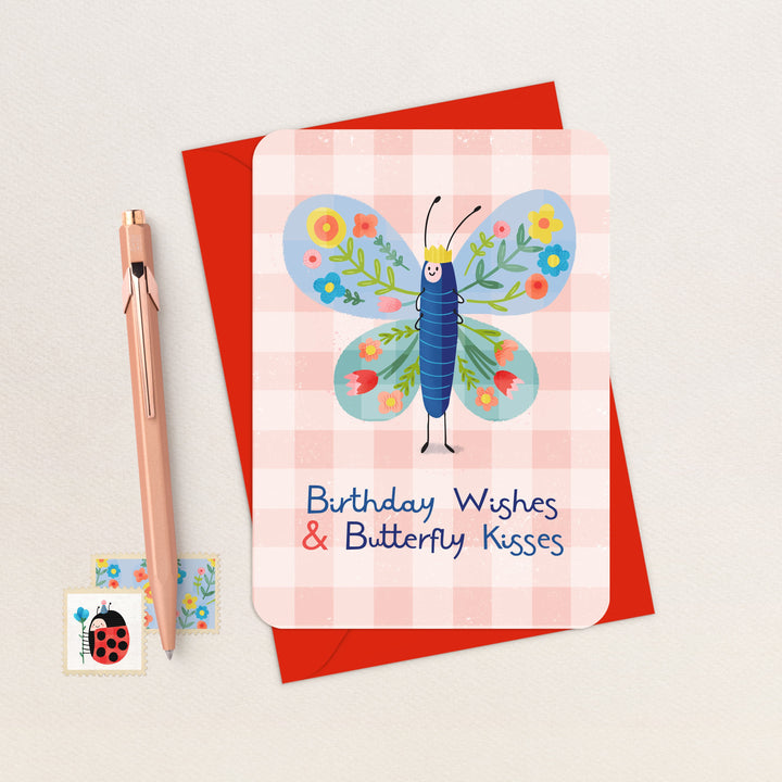 Birthday Wishes and Butterfly Kisses illustrated butterfly birthday card with red envelope, flat lay with pen and stamps
