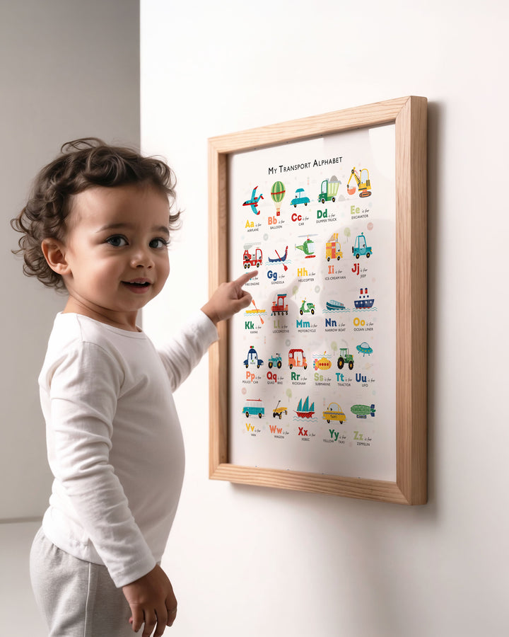 A toddler in a white long-sleeve shirt and light gray pants points at a framed alphabet poster on a white wall. The poster, titled &