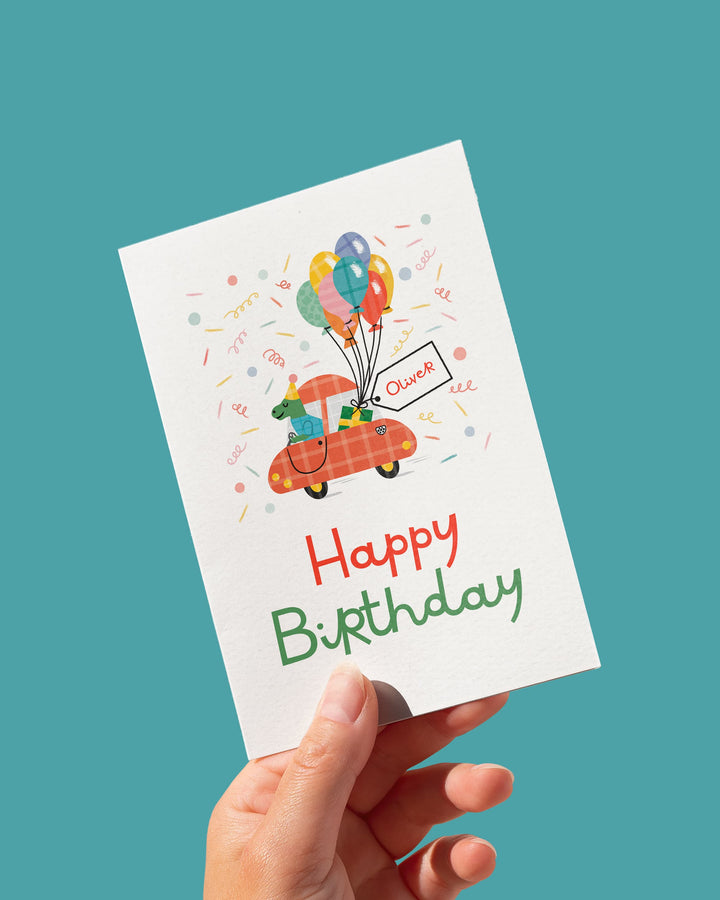 Hand holding a birthday card with a dinosaur in a car and colorful balloons and text on a teal background