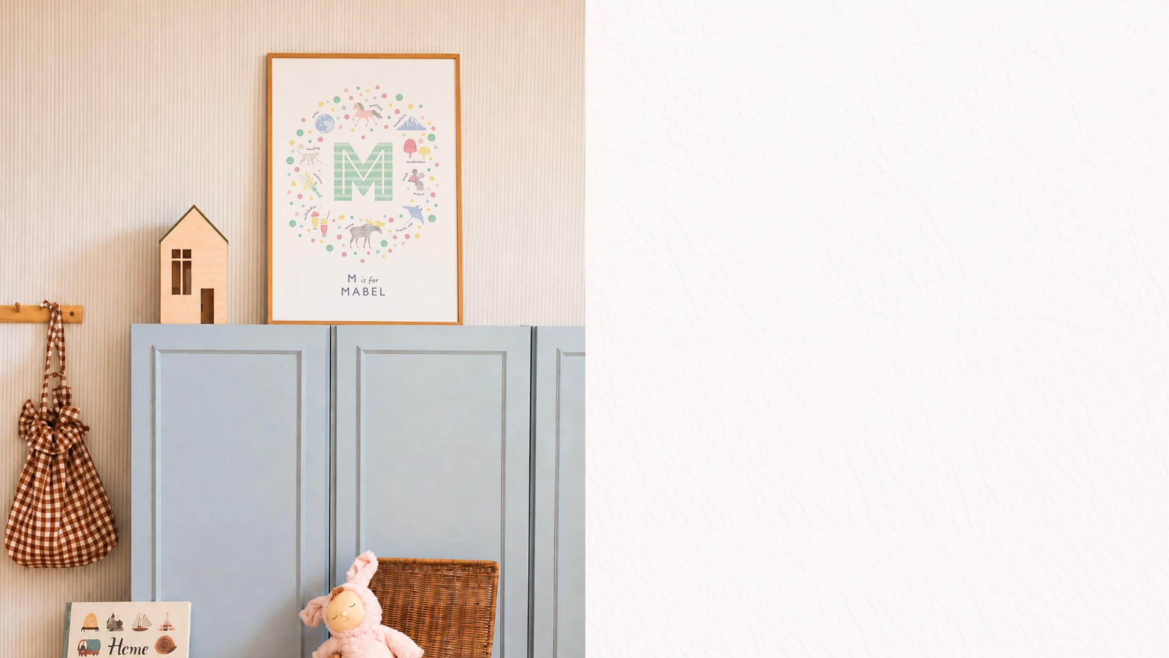 A framed personalized nursery art print featuring a colorful monogram 'M' and the name 'MABEL' on a light blue cabinet against a beige wall. The scene is styled with a small wooden house, a checkered bag, and a plush bunny.