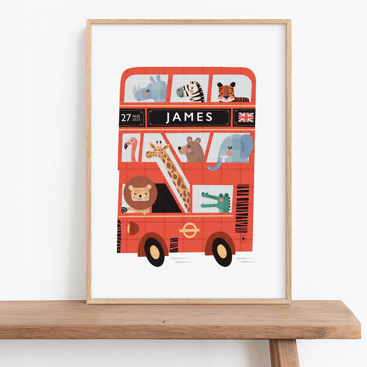 Personalised London Bus wall art in a natural wooden frame, sitting on a nursery wooden bench, featuring cute zoo animals like a lion, giraffe, and elephant. The custom name print also displays a birth date and flag of your choice.