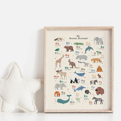 A framed Animal Alphabet print propped on a white surface next to a plush star-shaped pillow, showcasing a selection of illustrated animals from A to Z in a clean, minimalist style
