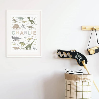 Personalised Dinosaur Name print framed in a white frame with mount on a magnolia wall in a playroom setting next to a hobby horse and play camera. 