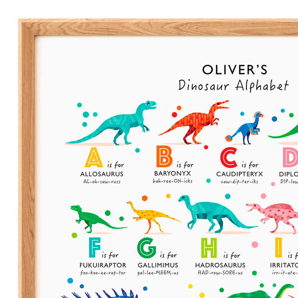 A framed children's poster titled 'Oliver's Dinosaur Alphabet' showing colorful illustrated dinosaurs labeled with letters and names, inside a light wood frame.