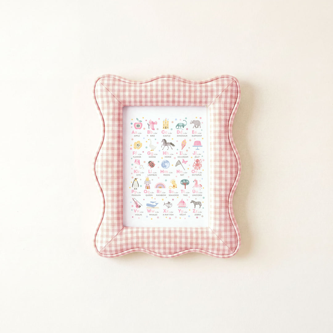 A scalloped decorative picture frame with a pink and white gingham border, featuring a colorful alphabet chart inside, shown against a plain off-white background.