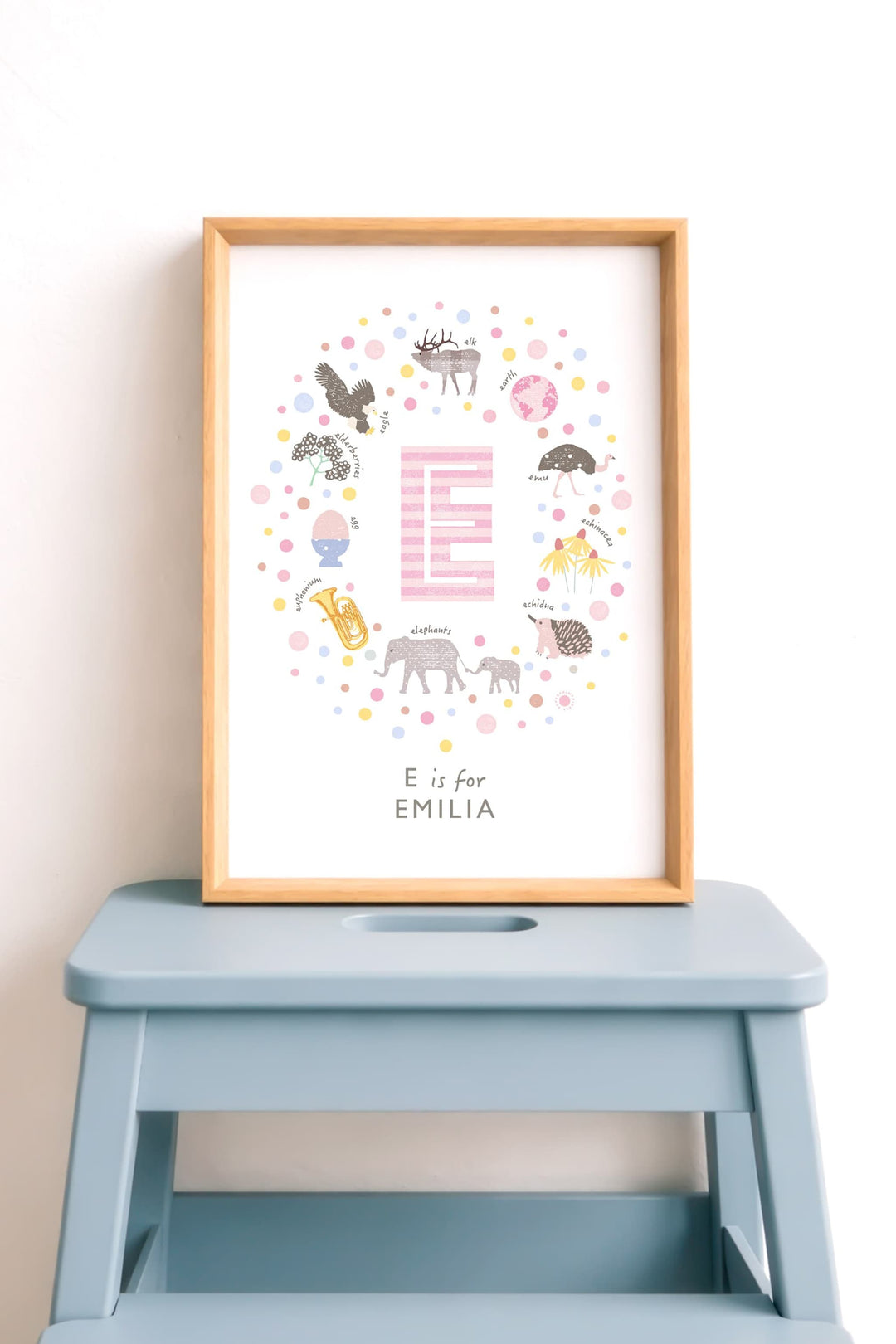 Pretty Framed personalised alphabet letter print with 'E is for EMILIA' on a blue step stool against a white wall.