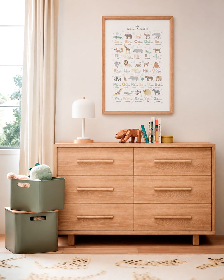 A framed animal alphabet chart hangs on a beige wall above a light oak six-drawer dresser in a styled nursery or children&
