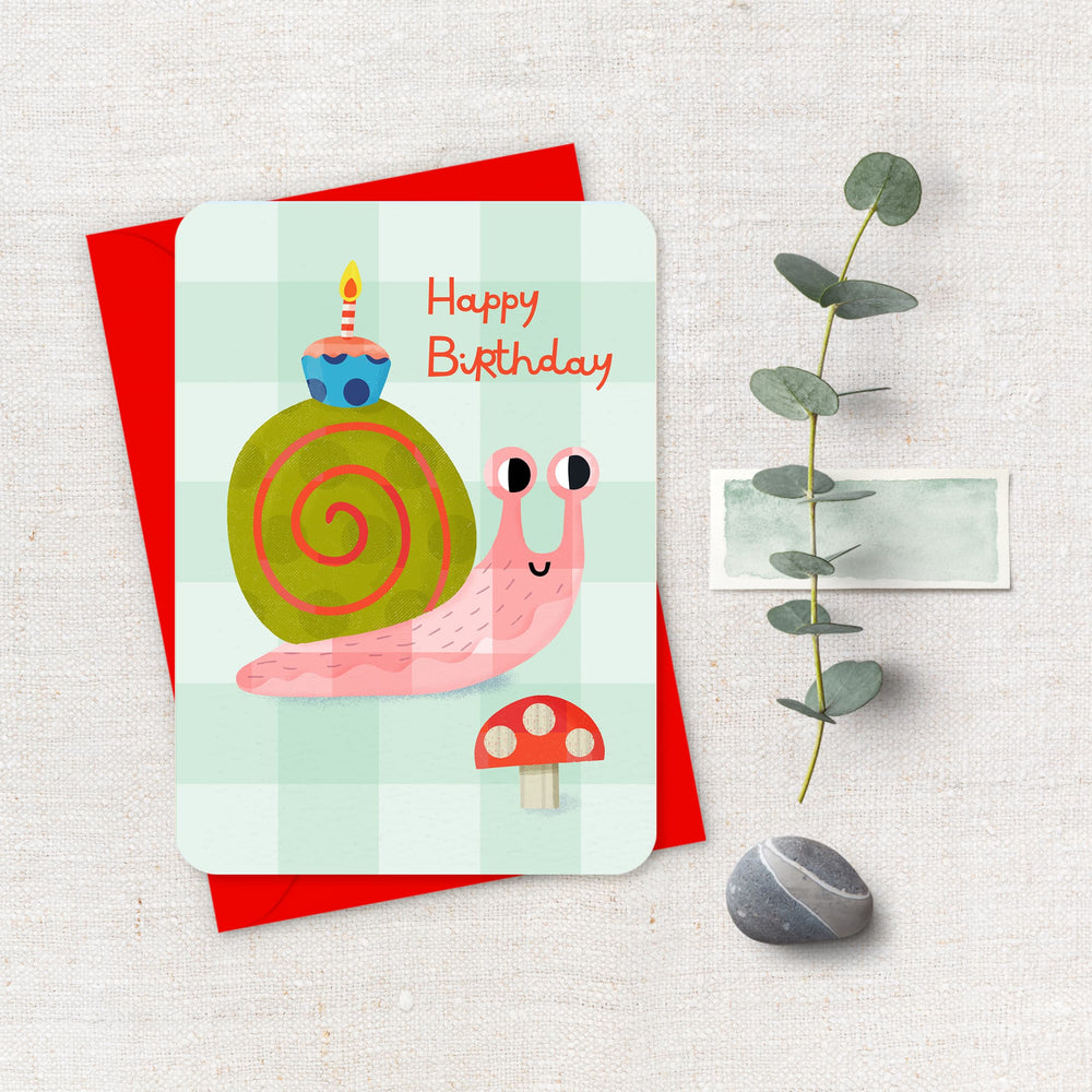 Birthday card with a snail illustration on a red envelope, accompanied by a plant and pebble.