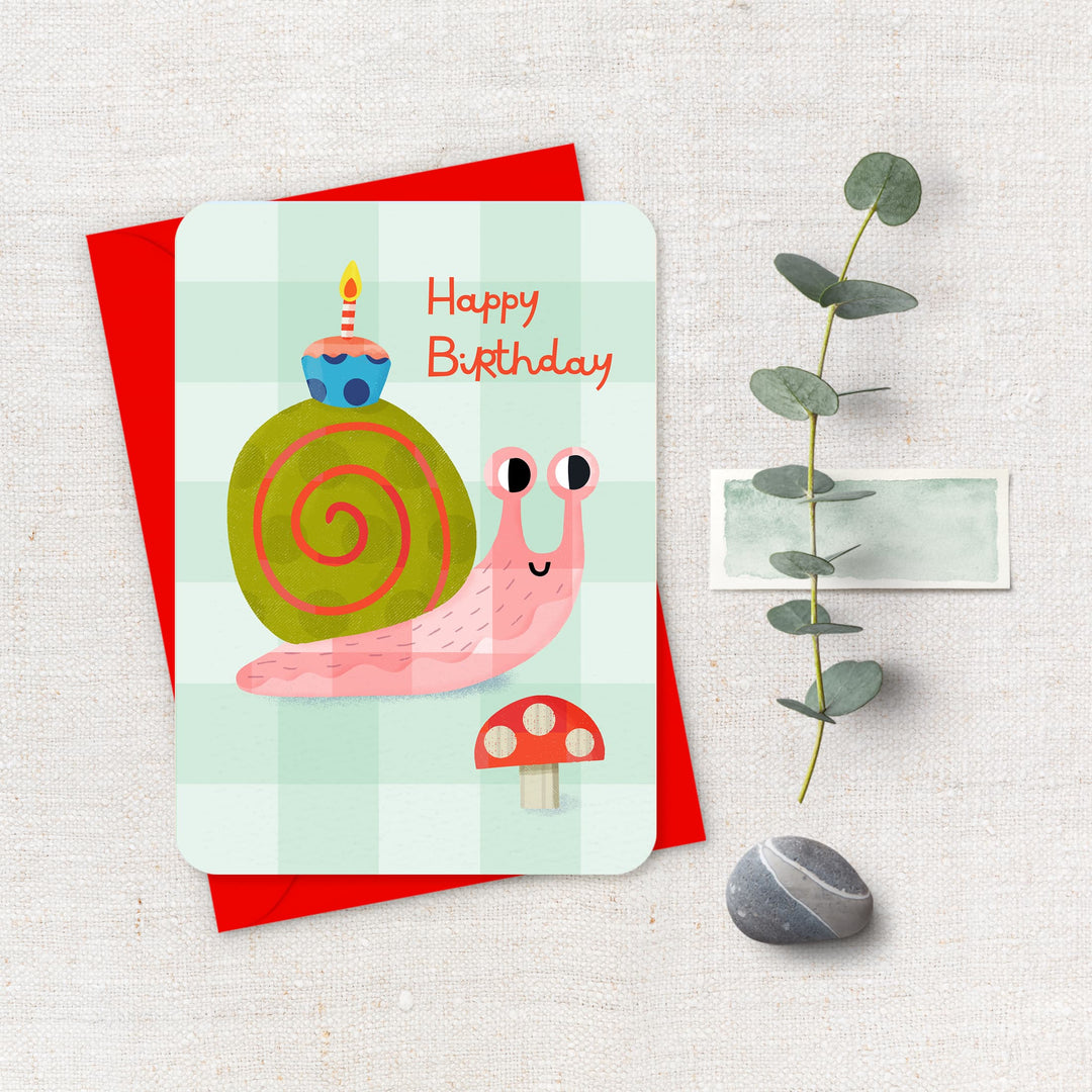 Birthday card with a snail illustration on a red envelope, accompanied by a plant and pebble.