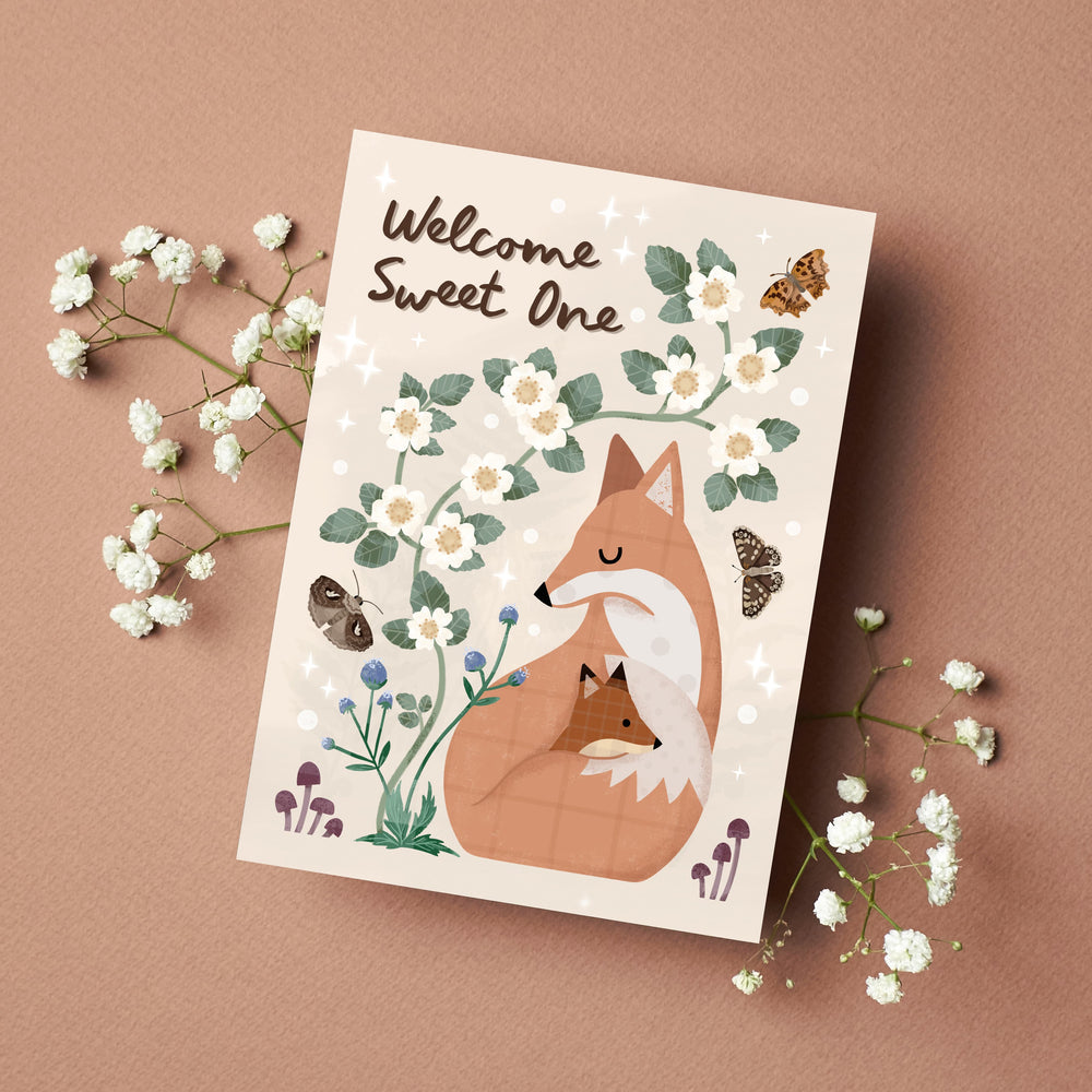 Illustrated woodland new baby card featuring a mother fox curled around her cub, surrounded by white blossom, butterflies and flowers. Welcome Sweet One lettering on a warm pink background with small white flowers surrounding the card