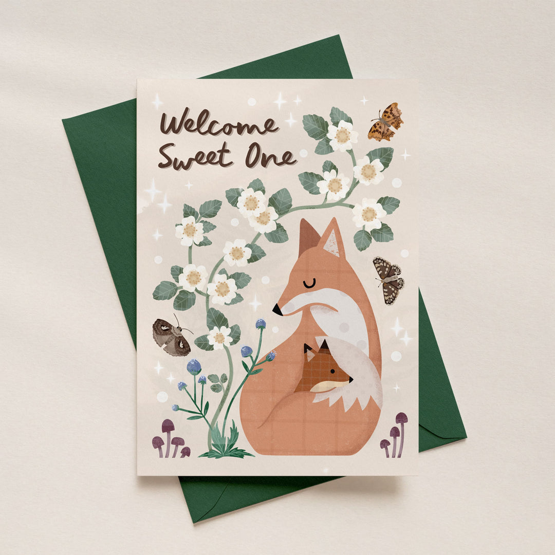 Woodland fox and cub new baby greeting card with Welcome Sweet One lettering, illustrated flowers and butterflies, displayed with its coordinating deep green envelope on a cream background.