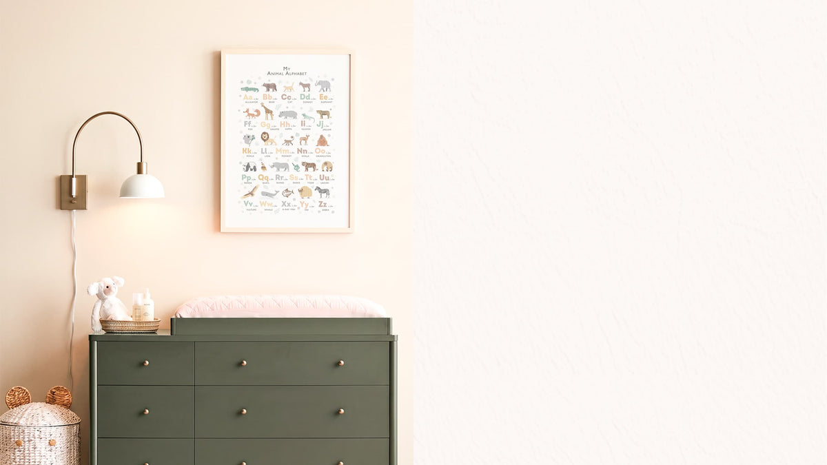 A framed animal learning chart hangs on a light beige wall above a dark green dresser in a styled nursery setting, with a wall lamp, changing pad, and small decor items.